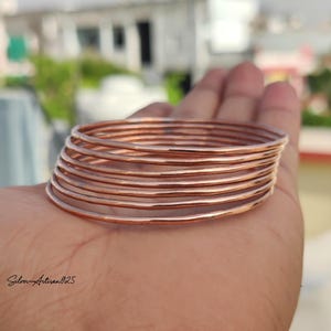 May include: A stack of thin, copper-colored bangles. The bangles are round and appear to be made of metal. The image is taken outdoors with a blurred background.
