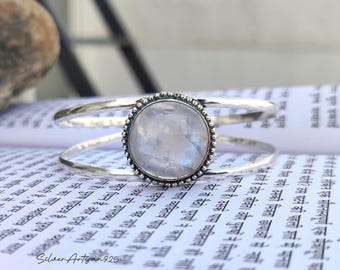 May include: A silver cuff bracelet with a large, round, iridescent moonstone center. The moonstone is set in a beaded bezel. The bracelet has a split design with two parallel bands. The background is a book with text.