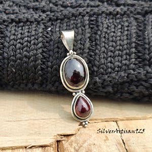 May include: A silver pendant featuring two red garnet gemstones. The top stone is oval, the bottom is teardrop-shaped. The pendant has a simple, elegant design. The text "SilverArtisan925" is visible on the bottom right.