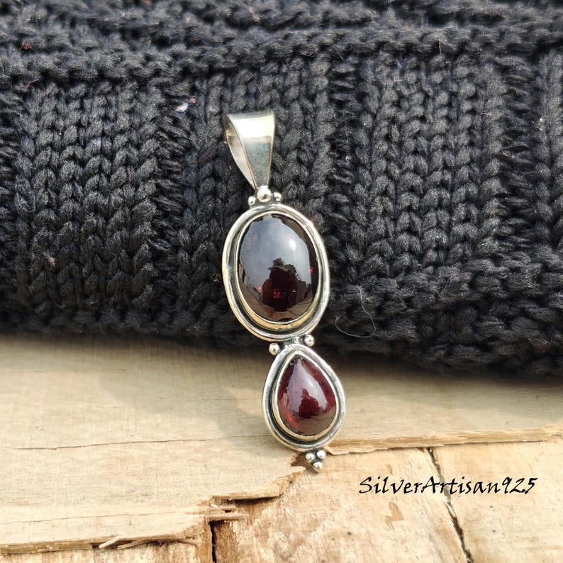 May include: A silver pendant featuring two red garnet gemstones. The top stone is oval, the bottom is teardrop-shaped. The pendant has a simple, elegant design. The text "SilverArtisan925" is visible on the bottom right.