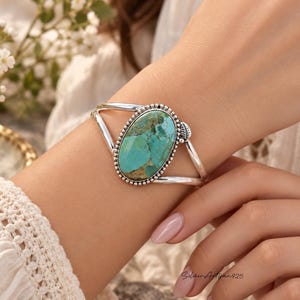 Könnte beinhalten: Ein Silberarmreif mit einem großen, ovalen Türkisstein mit brauner Matrix-Maserung. Der Stein ist in einer perlenbesetzten Silberfassung gefasst und weist an einer Seite ein kleines Blattdetail auf. Das Armband besteht aus zwei polierten Silberdrähten.