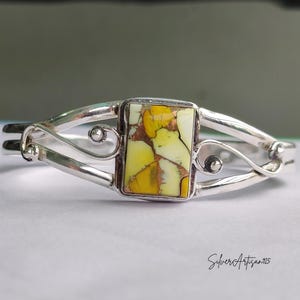 May include: A silver cuff bracelet with a rectangular, yellow and white stone centerpiece. The stone is set in a silver bezel and the bracelet has a decorative, open design with curved accents. The bracelet is a piece of jewelry.
