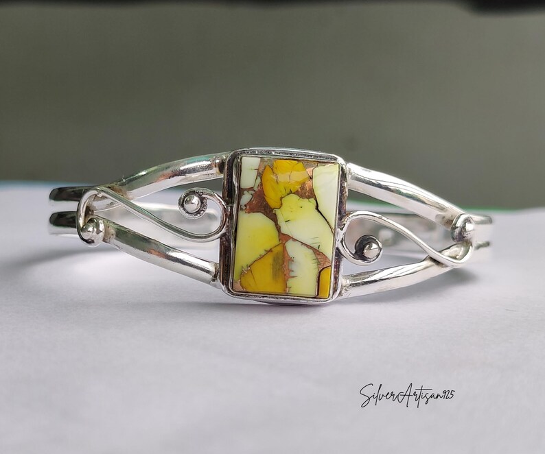 May include: A silver cuff bracelet with a rectangular, yellow and white stone centerpiece. The stone is set in a silver bezel and the bracelet has a decorative, open design with curved accents. The bracelet is a piece of jewelry.