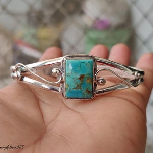 May include: A silver bracelet with a rectangular turquoise stone at the center. The turquoise stone has brown and blue-green hues. The bracelet has a decorative, open design with curved silver bands and small silver spheres. The text "Silver Artisan925" is in the bottom left corner.
