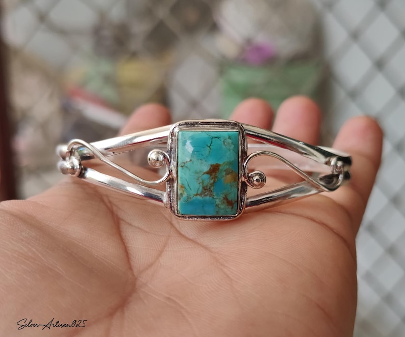 May include: A silver bracelet with a rectangular turquoise stone at the center. The turquoise stone has brown and blue-green hues. The bracelet has a decorative, open design with curved silver bands and small silver spheres. The text "Silver Artisan925" is in the bottom left corner.