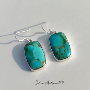May include: A pair of turquoise earrings with a rectangular shape and silver-colored metal trim. The earrings have a hook closure and a natural stone appearance, with shades of blue and brown.