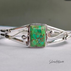 May include: A silver cuff bracelet with a rectangular, green turquoise stone at the center. The bracelet features a decorative, swirling design on the sides and is set against a neutral background. The artist's signature, "SilverArtisan925", is visible.