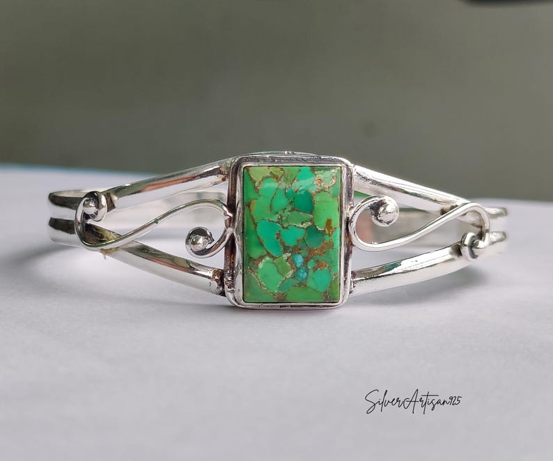 May include: A silver cuff bracelet with a rectangular, green turquoise stone at the center. The bracelet features a decorative, swirling design on the sides and is set against a neutral background. The artist's signature, "SilverArtisan925", is visible.