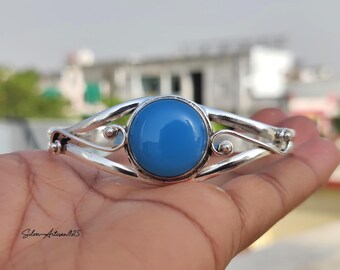 May include: A silver bracelet with a large, round, light blue stone at the center. The bracelet has a curved design with decorative swirls and small silver accents. The text "Silver Artisan 925" is visible.