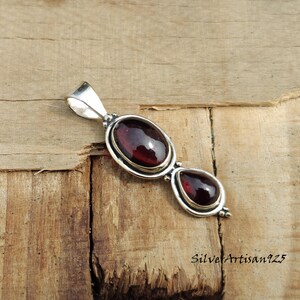May include: A silver pendant featuring two red-brown gemstones. The larger stone is oval, and the smaller one is teardrop-shaped. The pendant has a loop at the top for a chain and is set against a wooden background.