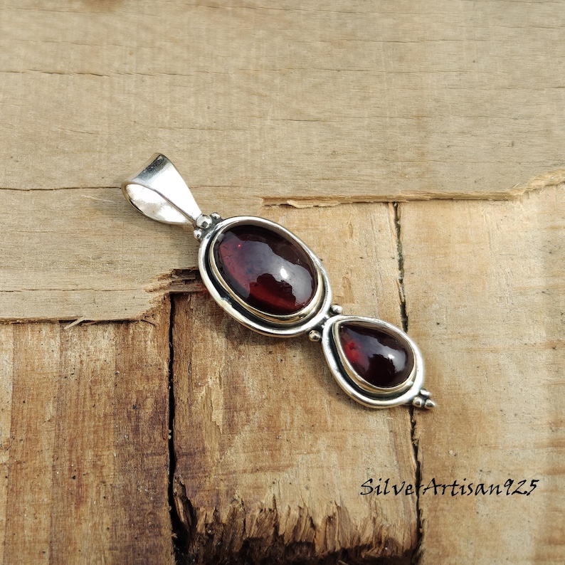 May include: A silver pendant featuring two red-brown gemstones. The larger stone is oval, and the smaller one is teardrop-shaped. The pendant has a loop at the top for a chain and is set against a wooden background.
