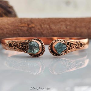 May include: A copper cuff bracelet featuring an open design with two raw, light blue-green stones set at each end. The metal band displays intricate embossed scrollwork and beaded detailing surrounding the stones. The text SilverArtisan925 is visible.