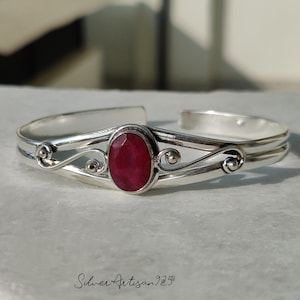 May include: A silver cuff bracelet featuring a large, oval, faceted ruby gemstone. The bracelet has a double-band design with decorative swirls and small silver beads. The text "Silver Artisan 925" is at the bottom.