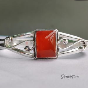 May include: A silver bracelet with a rectangular, vibrant orange gemstone at the center. The bracelet features a delicate, openwork design with curved lines and small decorative spheres. The inscription "SilverArtisan925" is visible.