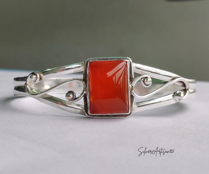 May include: A silver bracelet with a rectangular, vibrant orange gemstone at the center. The bracelet features a delicate, openwork design with curved lines and small decorative spheres. The inscription "SilverArtisan925" is visible.
