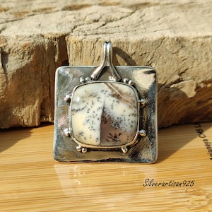 May include: A square silver pendant featuring a cream and brown stone with black inclusions. The stone is square-shaped and set within a square silver frame. The pendant has a loop for a chain. The text "Silverartisan925" is visible. The pendant is on a textured background.