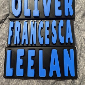 May include: Three black rectangular name plaques with raised blue lettering spelling "OLIVER", "FRANCESCA", and "LEELAN". The letters are bold and stand out against the black background.