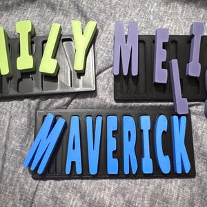 Puede incluir: Tres rompecabezas de nombres personalizados. Un rompecabezas deletrea "EMILY" en letras verdes, otro "MELISSA" en letras moradas, y el tercero "MAVERICK" en letras azules. Cada nombre está en una bandeja de rompecabezas negra.