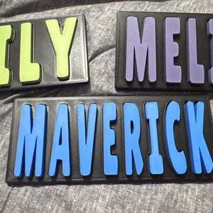 May include: Three wooden name puzzles. One puzzle spells "EMILY" in lime green letters, another spells "MELISSA" in purple letters, and the third spells "MAVERICK" in blue letters. Each name is on a black background.