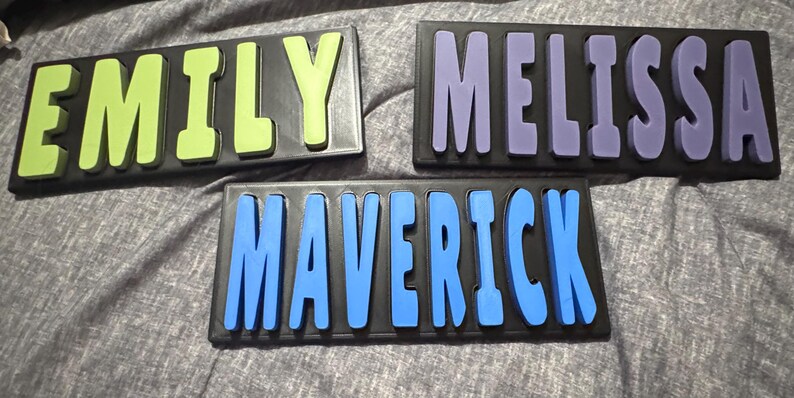 May include: Three wooden name puzzles. One puzzle spells "EMILY" in lime green letters, another spells "MELISSA" in purple letters, and the third spells "MAVERICK" in blue letters. Each name is on a black background.