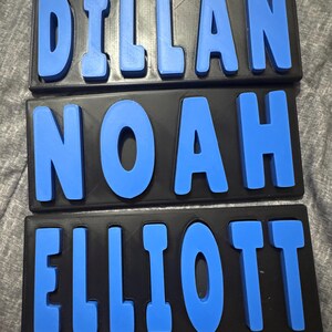 May include: Three personalized name plaques. Each plaque features a black base with raised, blue letters spelling out a name. The names are DILLAN, NOAH, and ELLIOTT. The letters are in a sans-serif font.