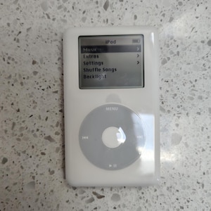 May include: A white iPod digital media player with a gray click wheel and a screen displaying the menu options: Music, Extras, Settings, Shuffle Songs, and Backlight. The iPod is on a speckled white surface.