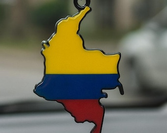May include: A car air freshener shaped like the map of Colombia, featuring the country's flag colors: yellow, blue, and red. The air freshener hangs from a black string attached to a car's rearview mirror.