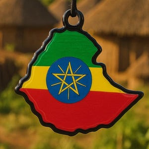 Puede incluir: Un colgante con la forma del mapa de Etiopía, con los colores de la bandera: verde, amarillo y rojo. Un círculo azul con una estrella amarilla en el centro. Cuelga de un cordón negro, con un fondo borroso de cabañas y montañas.