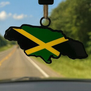 May include: A car air freshener shaped like the island of Jamaica, featuring the Jamaican flag's black, green, and yellow colors. The air freshener hangs from a string, with a road and trees visible in the background.