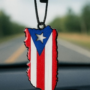 May include: A car air freshener shaped like the Puerto Rican flag, featuring red and white stripes, a blue triangle with a white star, and a black border. The air freshener hangs from a black cord.