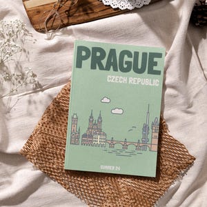 Könnte beinhalten: Ein grünes Reisetagebuch mit dem Text "PRAG TSCHECHISCHE REPUBLIK" und einer Skyline-Illustration der Stadt. Das Tagebuch trägt unten den Schriftzug "SUMMER 24".