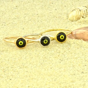 May include: Three gold rings with black and yellow evil eye charms on a sandy background.