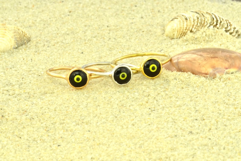 May include: Three gold rings with black and yellow evil eye charms on a sandy background.