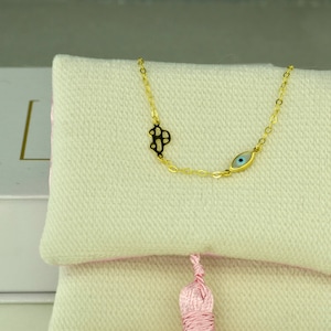 May include: A gold-colored necklace with a cross and an eye charm. The necklace is displayed on a white cushion with a pink tassel. The cross charm is black and gold, and the eye charm has blue and white details.