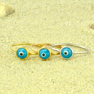 May include: Three gold rings with blue evil eye charms. The rings are on a sandy surface.