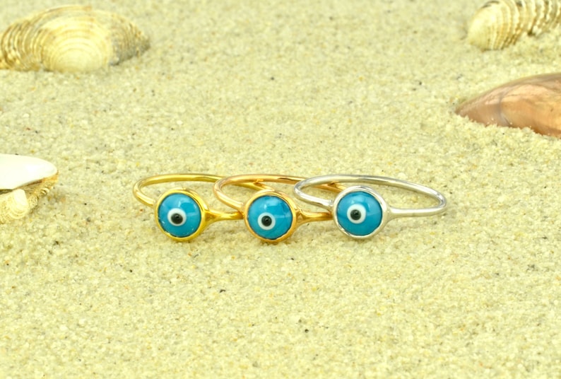 May include: Three gold rings with blue evil eye charms. The rings are on a sandy surface.