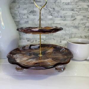 May include: Two-tiered serving tray with a brown, white, and gold marbled resin finish. The tray has a scalloped edge and sits on clear acrylic feet.