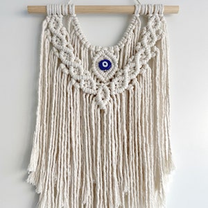 May include: A white macrame wall hanging with a blue evil eye charm in the center. The hanging is made with cotton cord and has a wooden dowel at the top.