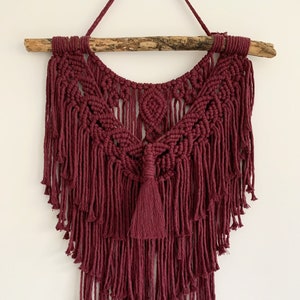 May include: A burgundy macrame wall hanging with a wooden dowel at the top and a tassel at the bottom. The wall hanging is made with a variety of knots and has a fringe design.