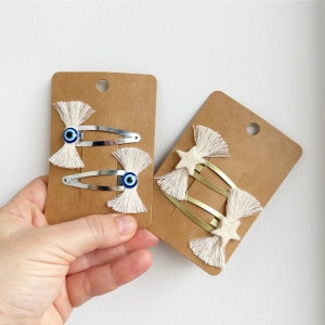 May include: Two sets of hair clips with white tassels. One set features blue evil eye charms and the other set features gold star charms.