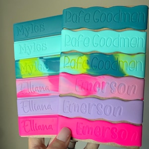 May include: A collection of rectangular silicone bands with wavy edges, featuring embossed names including Myles, Rafe Goodmen, Elliana, and Emerson. The bands come in various solid colors and marbled patterns, including teal, mint, neon pink, and lavender.