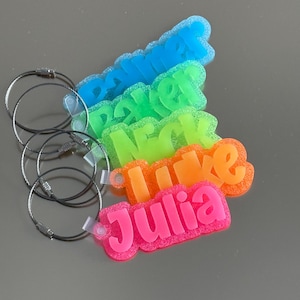 May include: A collection of colorful name tags with attached metal key rings. The names are in various colors: blue, green, orange, and pink. The name "Julia" is visible.