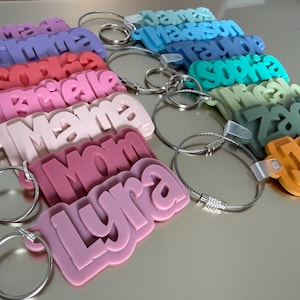 May include: A collection of colorful, personalized keychains. Each keychain features a name in a layered, 3D design. The names are in various colors, including purple, red, pink, blue, green, and orange, with silver key rings.