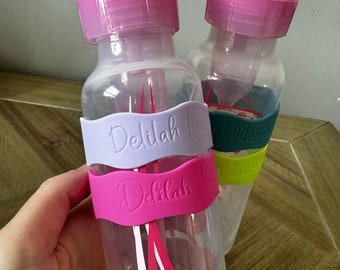 May include: Two clear plastic baby bottles with pink lids and silicone sleeves. The sleeves have the name "Delilah" written on them.