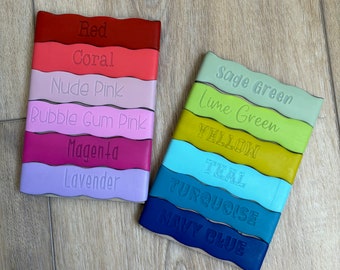 May include: A set of 12 leather bookmarks in various colors, including red, coral, nude pink, bubble gum pink, magenta, lavender, sage green, lime green, yellow, teal, turquoise, and navy blue. Each bookmark has a wavy edge and the color name is embossed on the bookmark.