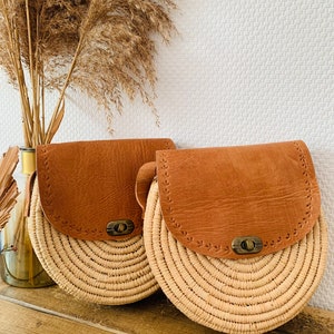 May include: Two round straw bags with brown leather flaps and metal closures. The bags have a woven design and are sitting on a wooden surface.
