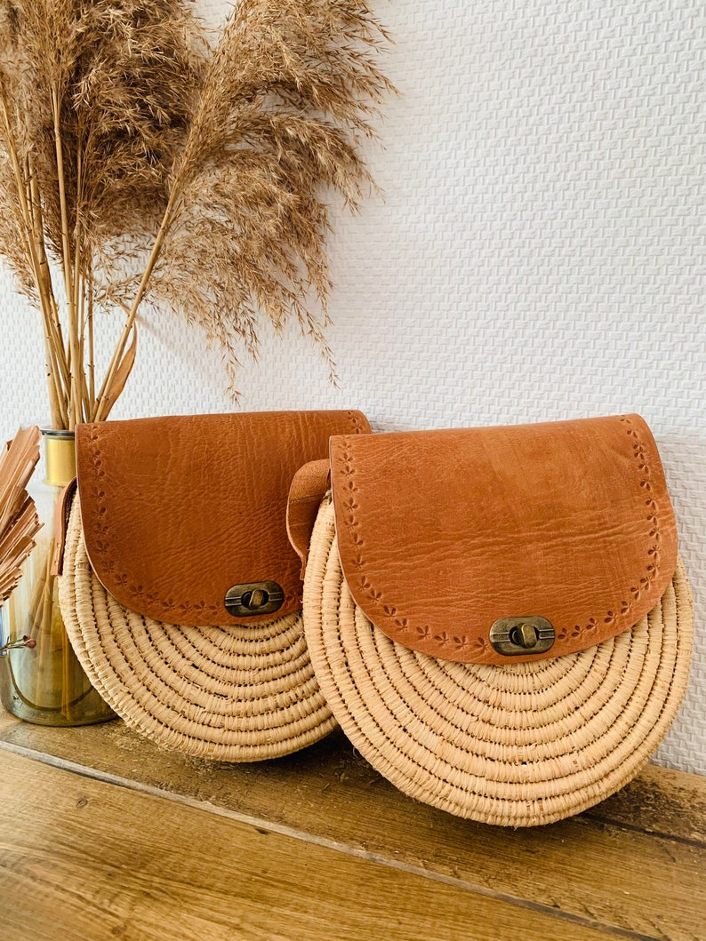 May include: Two round straw bags with brown leather flaps and metal closures. The bags have a woven design and are sitting on a wooden surface.