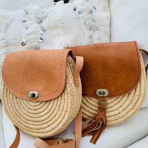 May include: Two round straw bags with leather accents. The bags have a brown leather flap with a metal buckle and a long brown leather strap. One bag has a tassel.