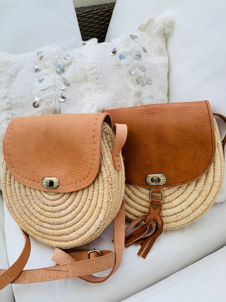 May include: Two round straw bags with leather accents. The bags have a brown leather flap with a metal buckle and a long brown leather strap. One bag has a tassel.
