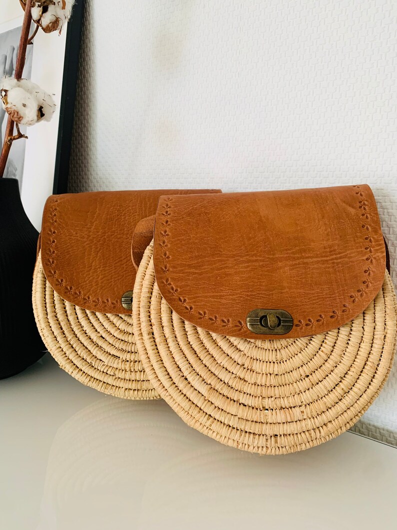 May include: Two round straw bags with brown leather flaps and a metal clasp. The bags have a woven design and are perfect for summer.